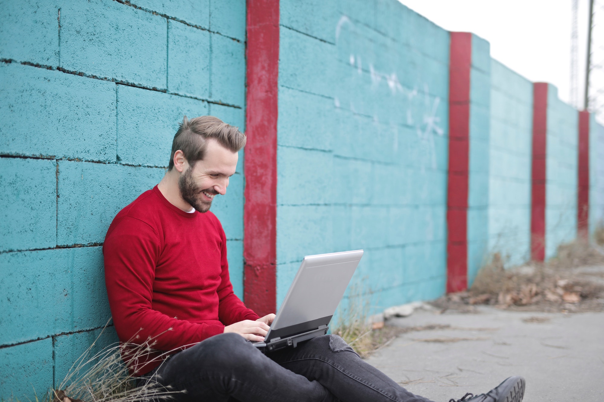 man leans against a blue wall outside and works at his laptop. he wears a long sleeved red shirt and is smiling down at the laptop