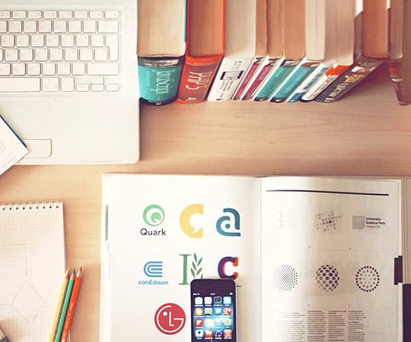 an open book with an iphone on top of it. the desk it's on also has a stack of books behind the open book and a laptop next to the stack