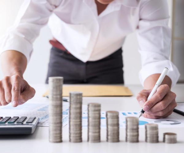 person hunched over a desk with a pen in their hand writing on a piece of paper. a stack of coins sits in front of them in ascending size.