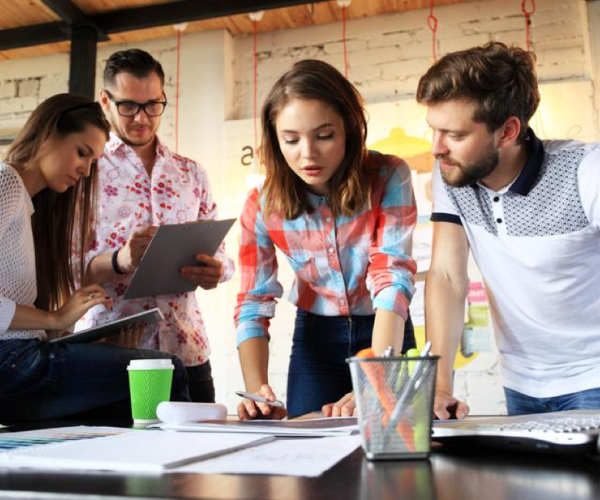 a group of 4 people are hunched over a desk with paper and office supplies on it