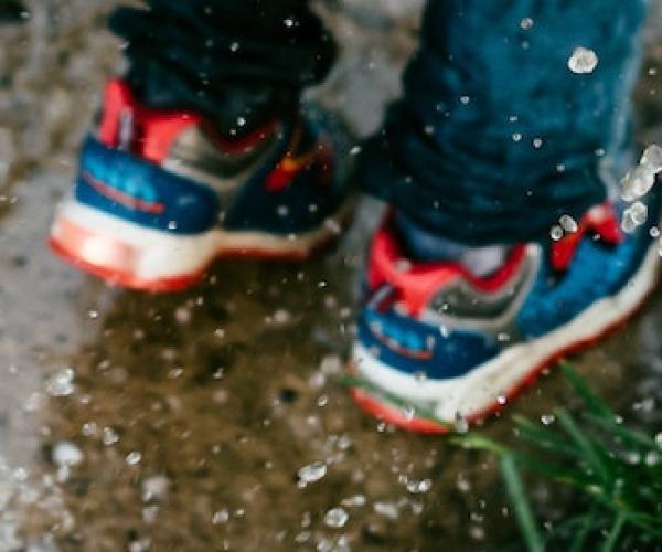 a child wearing blue and red sneakers splashes in a puddle. there is a grassy patch to their right and there are white pebbles to their left