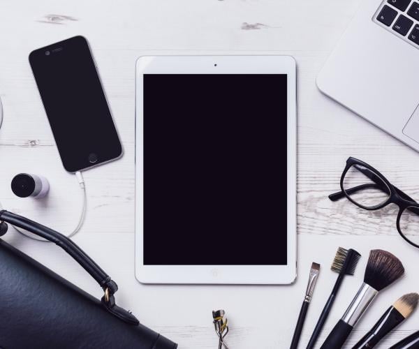 bird's eye view photo of a white desk with white electronics on top: an ipad, iphone, black pair of glasses, and some pens and paint brushes