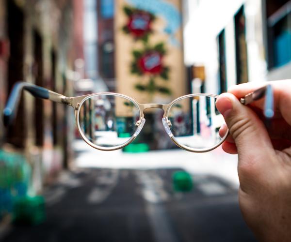 a hand holding out a pair of round glasses as the camera sees through them. the background is a city street