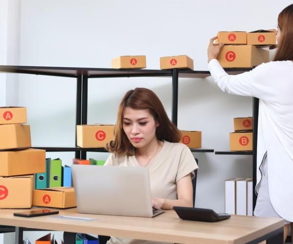 2 women in an office, one is sitting facing the viewer at a desk on a laptop, numerous boxes are on the desk and around the room. the 2nd woman's back is facing the viewer and she is adjusting one of the boxes behind the 1st woman