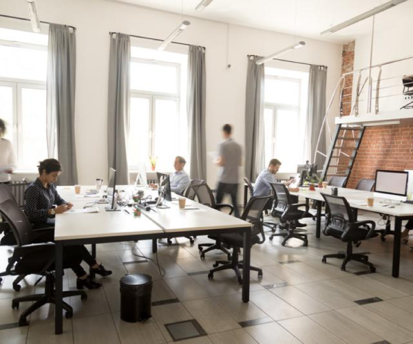 an office with many people in it sitting at desks working on computers or walking around. the office is very modern-looking and has wide floor-to-ceiling windows