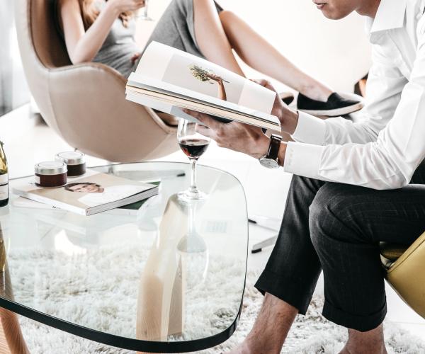 person sitting reading a book at a table with another person sitting further away in a chair also reading. both of their heads are out of the shot