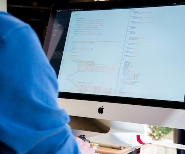 a person in a blue shirt sits in front of an imac computer that has code on the screen