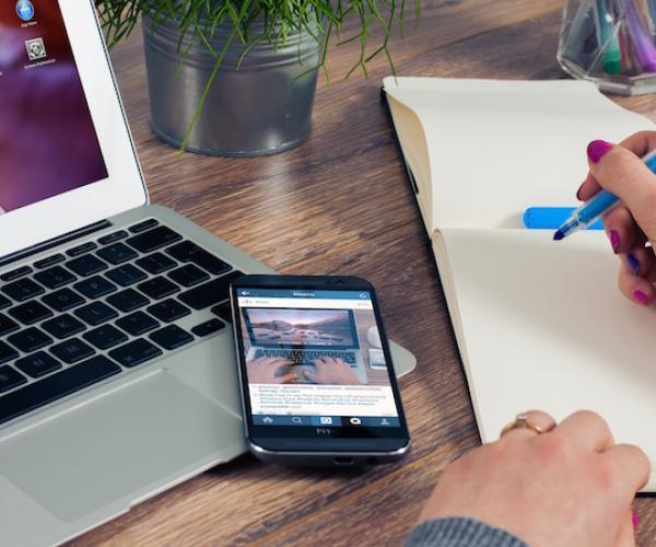 a person taking notes in an empty notepad with a blue marker. to their left is a phone resting on top of a laptop