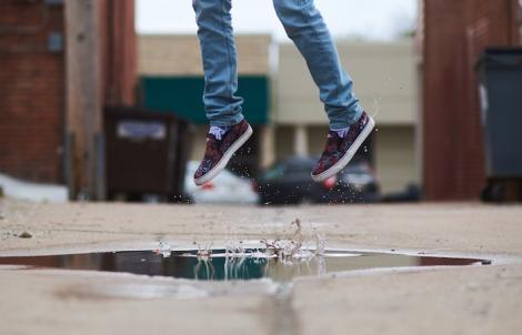 a person in lue skinny jeans and black worn vans shoes jumps over a puddle