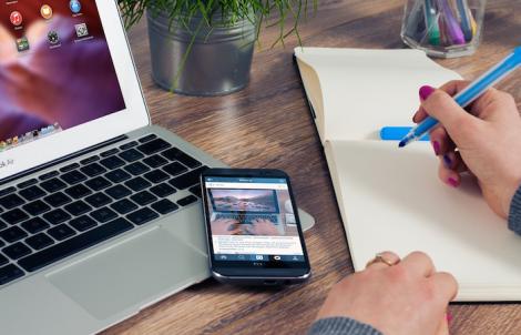 a person taking notes in an empty notepad with a blue marker. to their left is a phone resting on top of a laptop