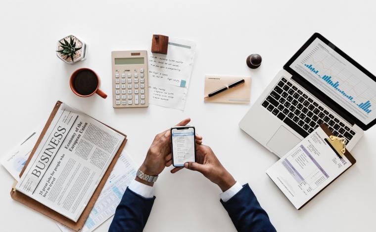 a person's hands holding a phone with other office items laid out around them on a white desk