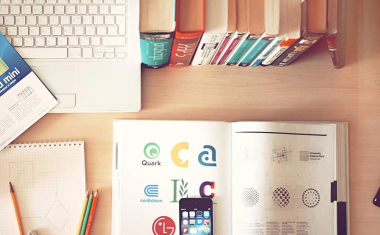 an open book with an iphone on top of it. the desk it's on also has a stack of books behind the open book and a laptop next to the stack