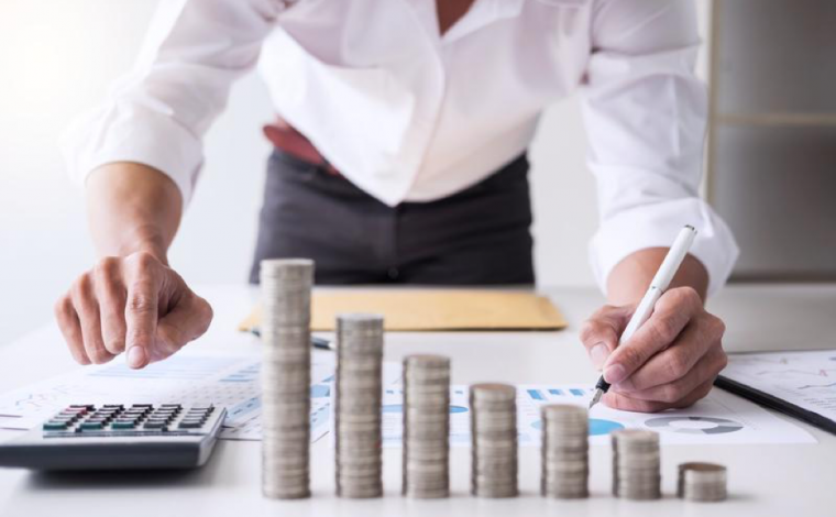 person hunched over a desk with a pen in their hand writing on a piece of paper. a stack of coins sits in front of them in ascending size.