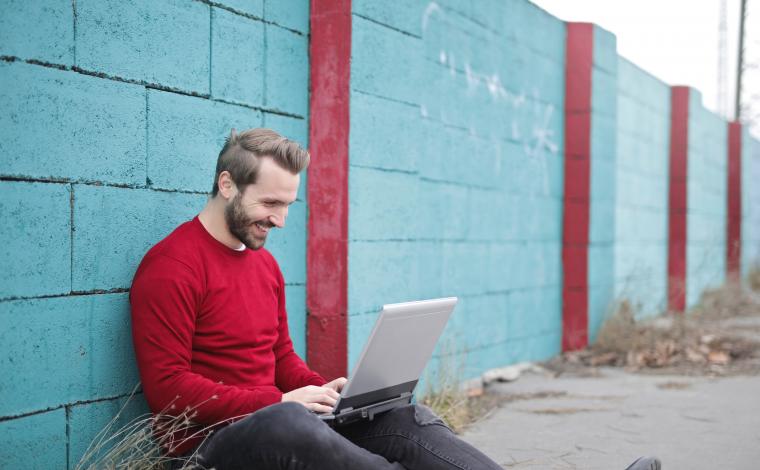 a man with a red long sleeved shirt sitting next to a blue wall on his laptop