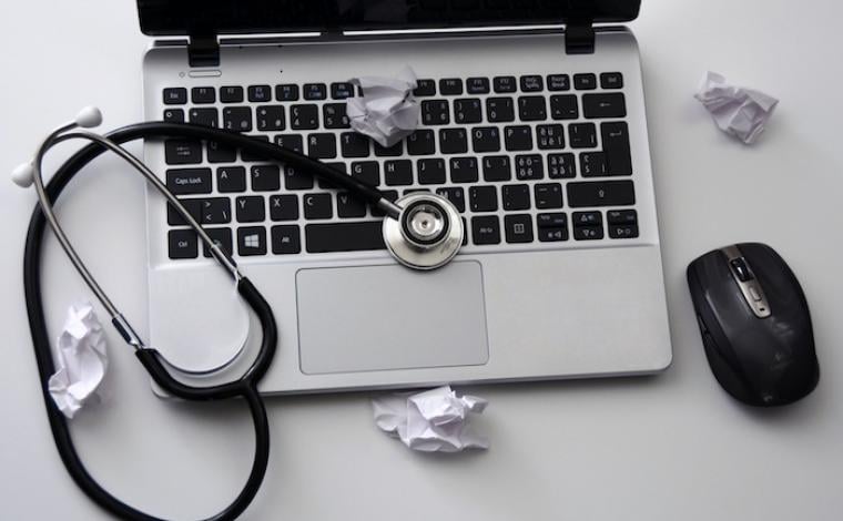 an open laptop with a stethoscope on top of it. a wireless mouse sits to the right of the laptop and crumpled white pieces of paper are around the laptop