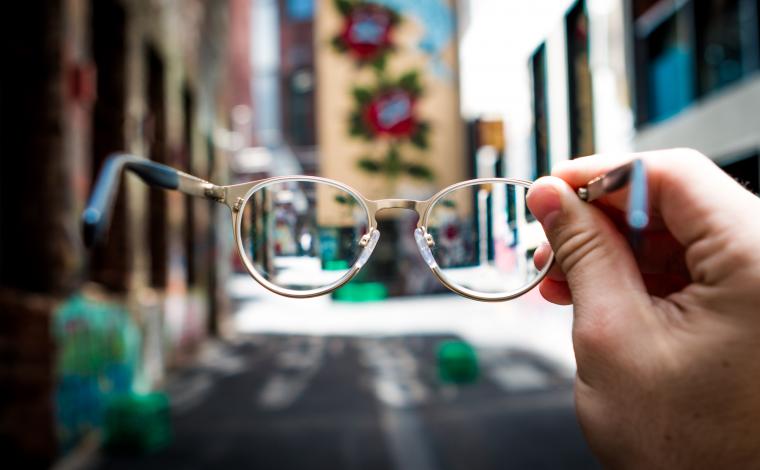 a hand holding out a pair of round glasses as the camera sees through them. the background is a city street