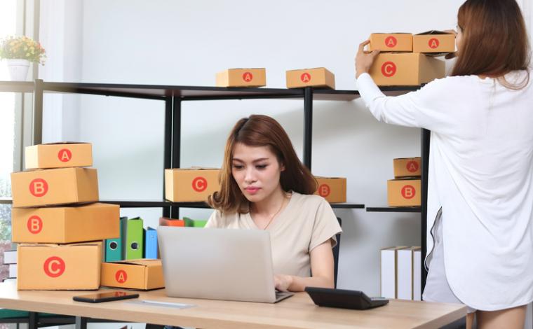 2 women in an office, one is sitting facing the viewer at a desk on a laptop, numerous boxes are on the desk and around the room. the 2nd woman's back is facing the viewer and she is adjusting one of the boxes behind the 1st woman
