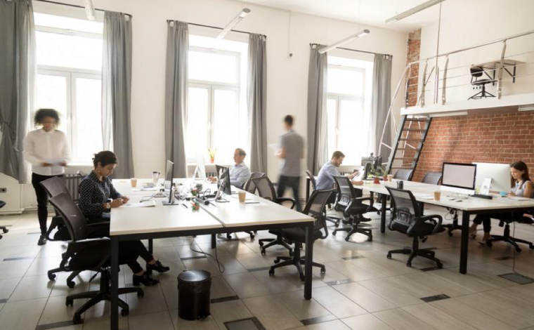 an office with many people in it sitting at desks working on computers or walking around. the office is very modern-looking and has wide floor-to-ceiling windows