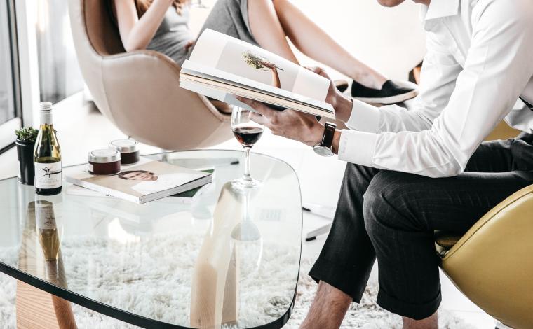 person sitting reading a book at a table with another person sitting further away in a chair also reading. both of their heads are out of the shot