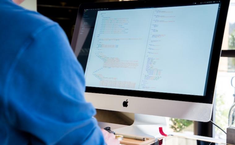 a person in a blue shirt sits in front of an imac computer that has code on the screen