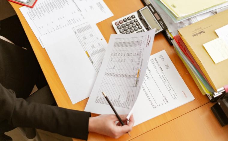 a brightly colored wooden desk with a person sitting at it taking notes with a few pieces of paper in front of them on the desk. they hold one of the papers and a pen in their right hand