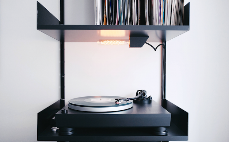 a record player with a record on it. the player sits on a piece of furniture that is the same width as the player, and has a top section with shelving that houses various records.