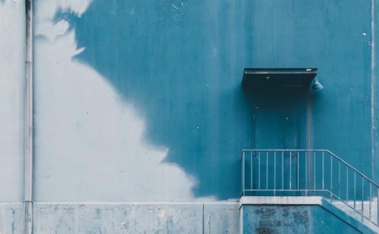 the side of a half blue painted building. there's a raised platform lining the building with a little railing and door on the right side