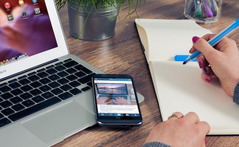 a person taking notes in an empty notepad with a blue marker. to their left is a phone resting on top of a laptop